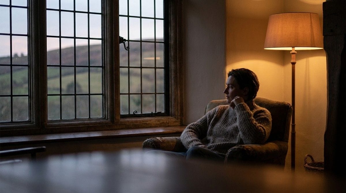 Person sitting in an armchair and looking thoughtfully out a window in a warmly lit room.