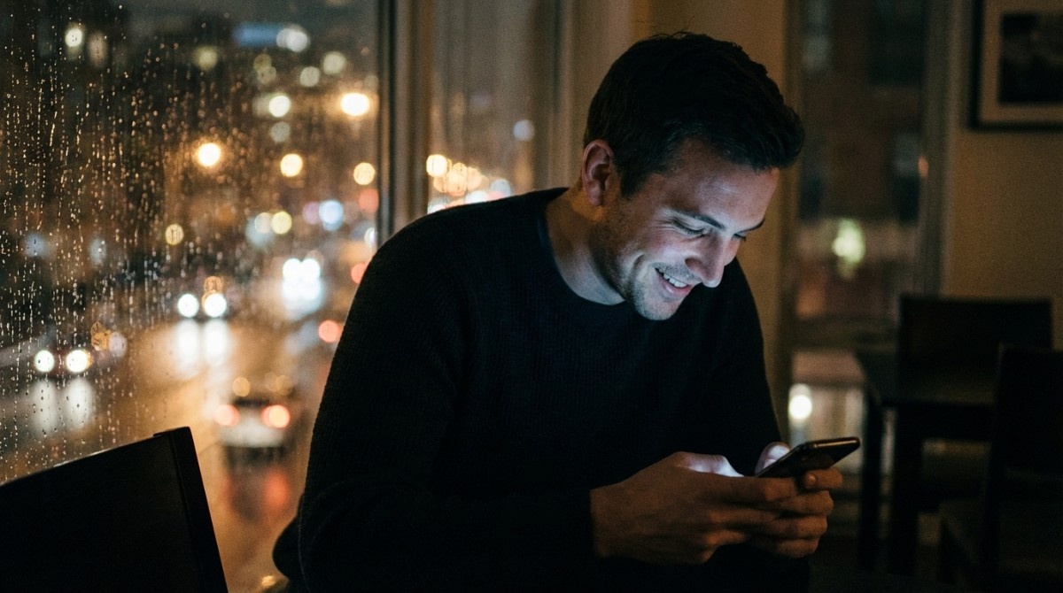 Man sitting by a window at night smiling at his phone with city lights blurred in the background